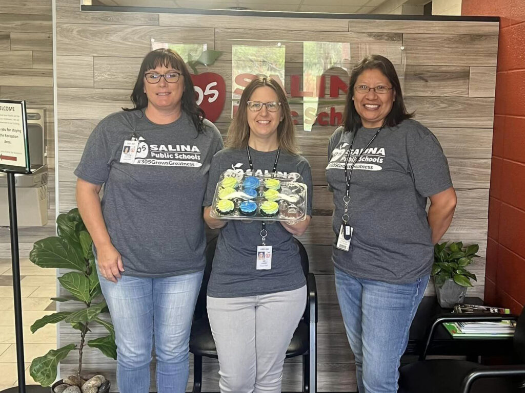 Three people are standing in front of a sign that reads "Salina Public Schools." They are all wearing matching gray t-shirts with the school's name and logo. The person in the middle is holding a tray of cupcakes with yellow and blue frosting.