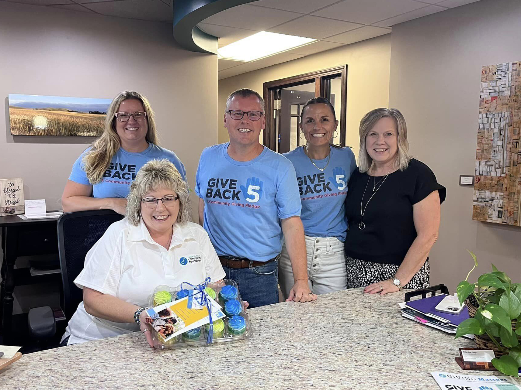 A group of five adults stand around a reception desk. Four are standing, and one is seated while holding a gift basket. Three of them are wearing light blue "Give Back 5" T-shirts, while the other two are wearing black tops.