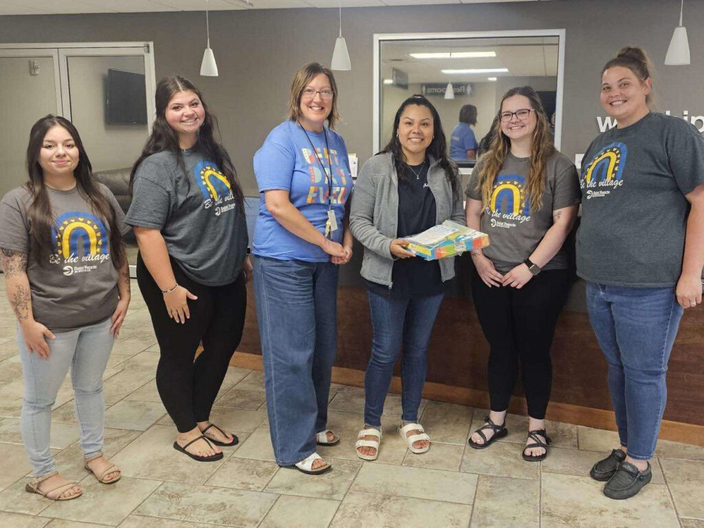 A group of six women stands together in an office setting. Two of them are holding a pack of colored markers. They are smiling and appear to be part of a work team or event. Some are wearing T-shirts with a rainbow logo and text that reads "Let the Village Donate.