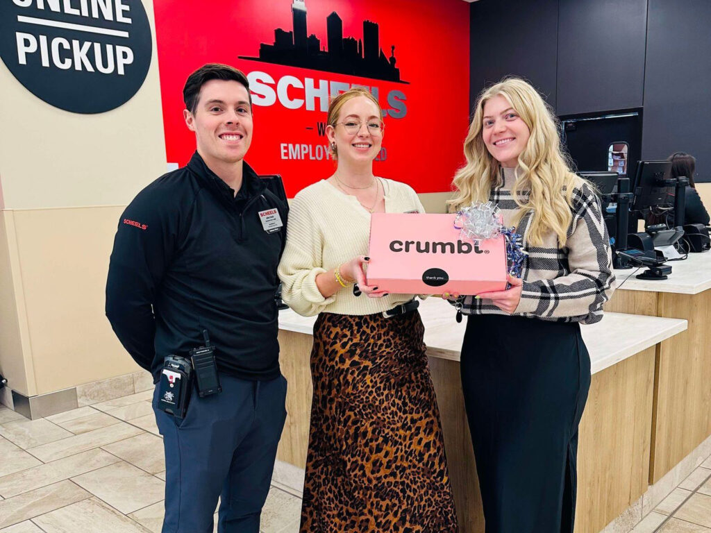 Three people stand indoors at Scheels, near the online pickup counter. The person in the middle holds a pink Crumbl Cookies box, and all three are smiling. The background features store branding in red, black, and beige colors.