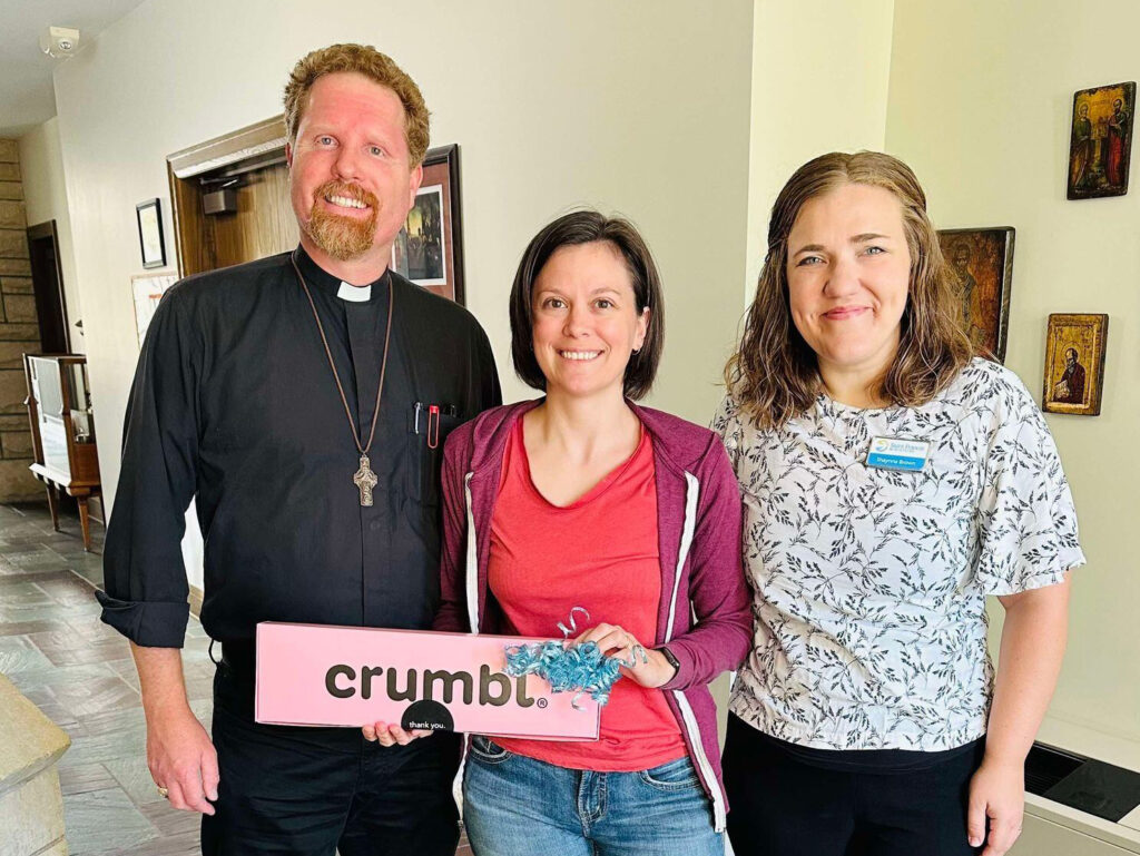 Three people, two women and a man, are standing indoors. The man on the left is wearing a clerical shirt with a cross necklace. The woman in the middle is holding a pink box labeled "Crumbl," and the woman on the right is wearing a floral shirt and a name tag.