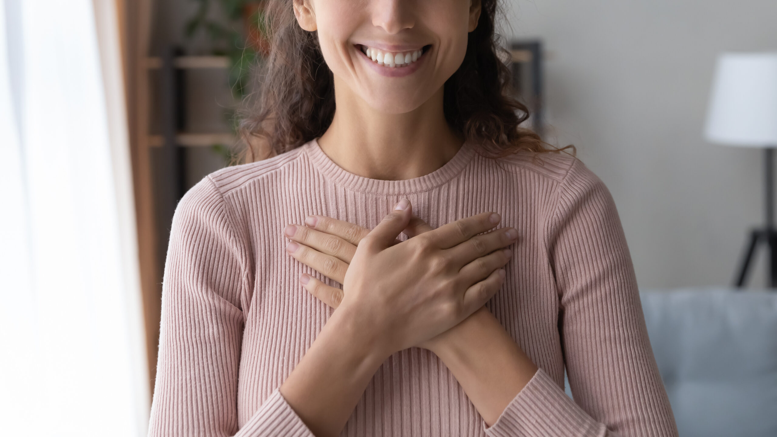 A person is standing indoors, smiling with their hands crossed over their chest. They are wearing a light pink, long-sleeved ribbed top. The background is blurred, showing some furniture and plant.