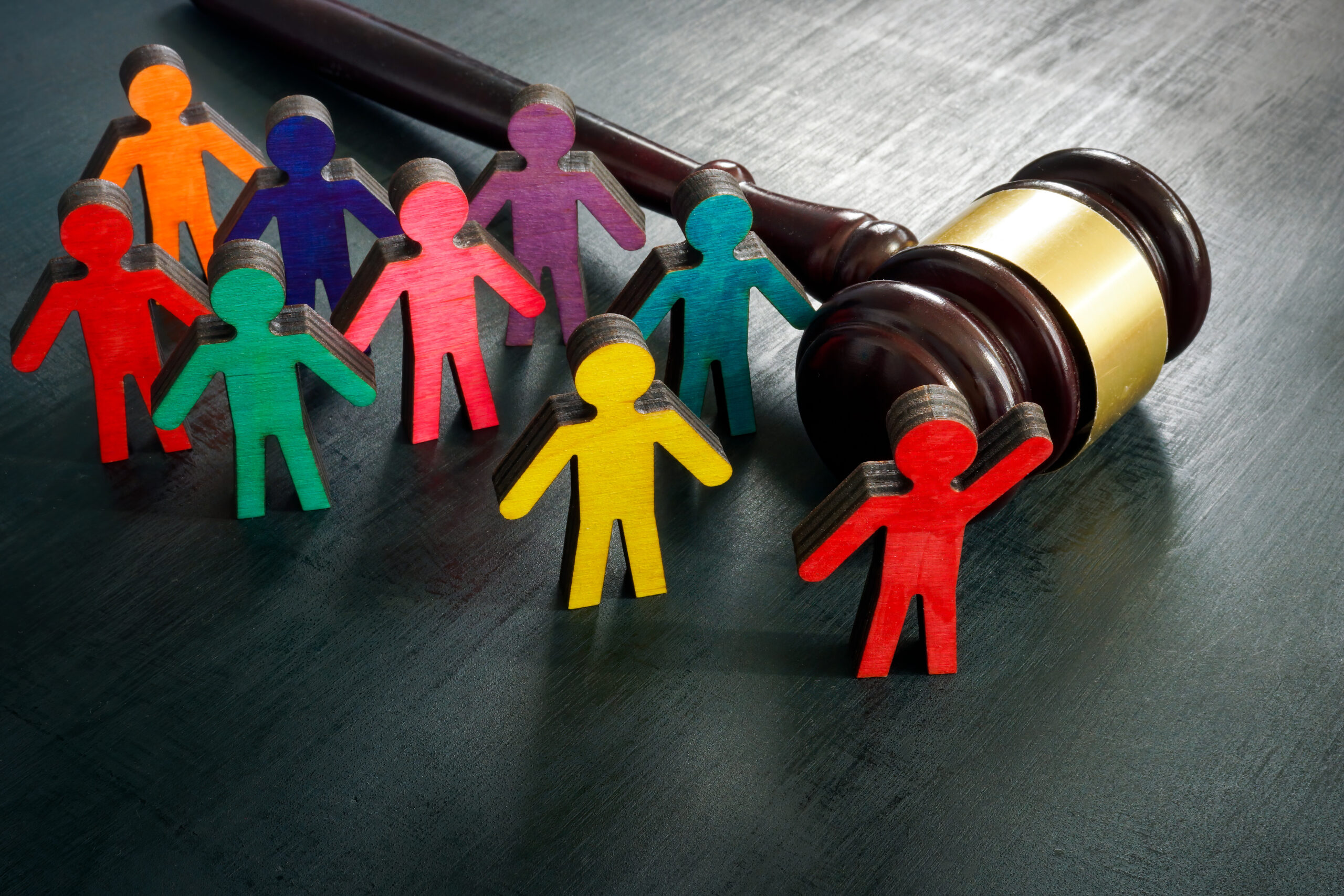 A wooden gavel lies on a dark surface next to several colorful cut-out figures in various colors, including red, orange, yellow, and green. The figures appear to represent people, standing in a semi-circle around the gavel.