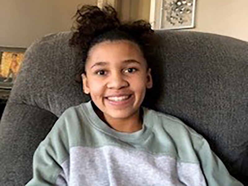 A person with curly hair tied up is smiling at the camera. They are sitting on a cushioned chair and wearing a gray and green top. The background shows part of a room with a framed picture on the wall.