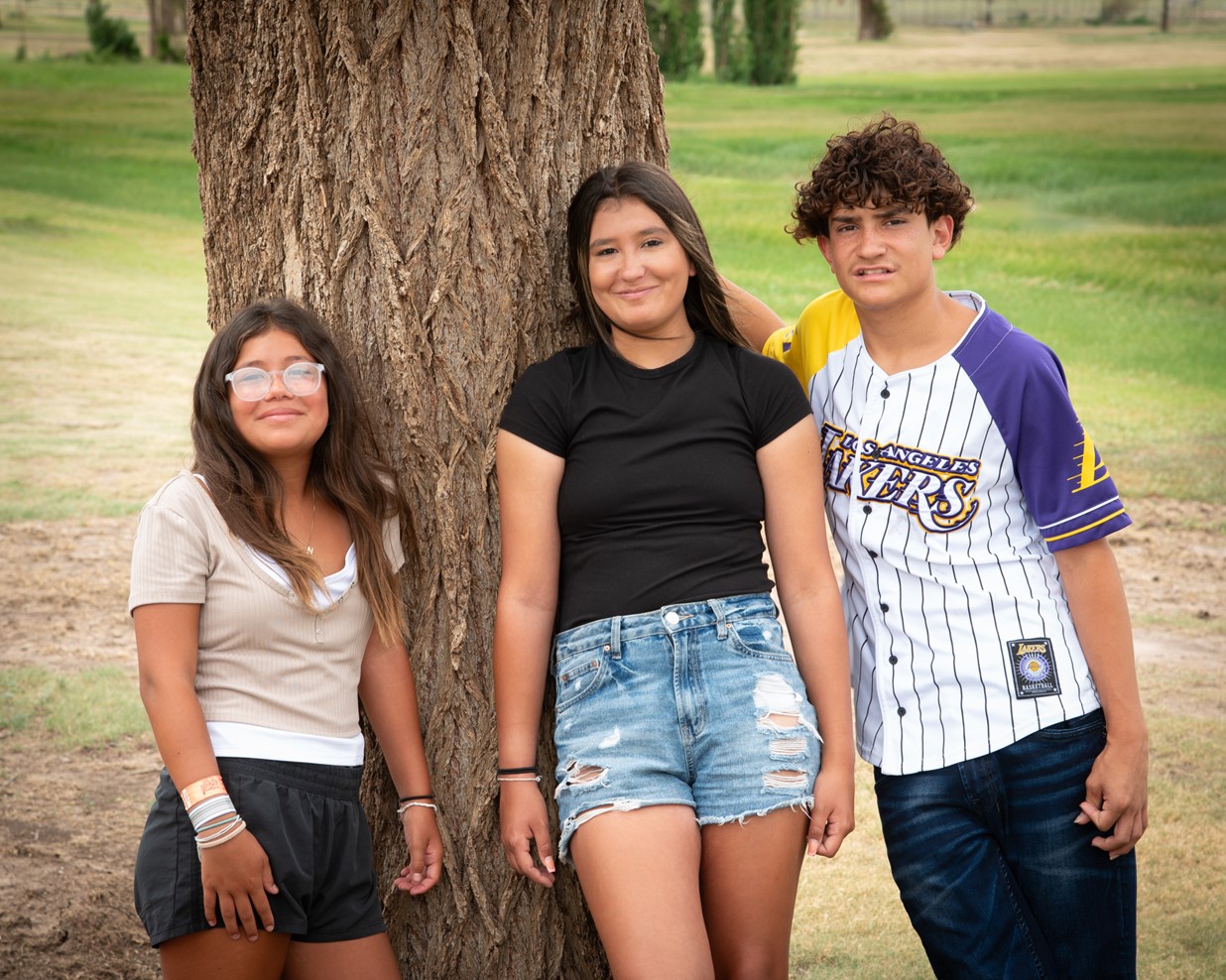 Three teenagers stand outdoors next to a tree. The girl on the left wears glasses, a beige top, and black shorts. The middle girl has on a black top and denim shorts. The boy on the right is in a Los Angeles Lakers jersey and jeans. A grassy field is in the background.