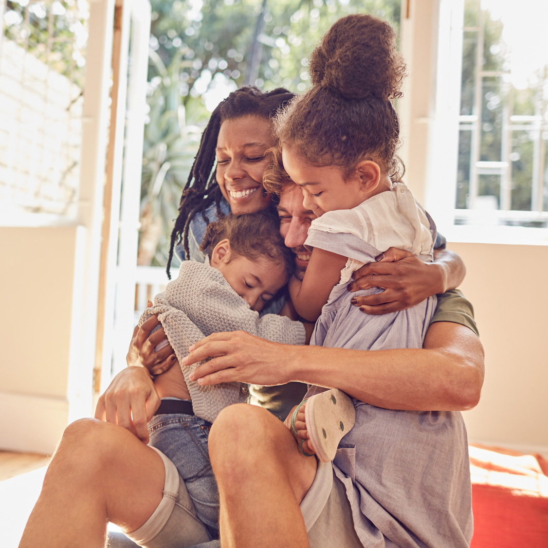 A family of four sits closely, hugging each other. The woman and the man are seated on the floor, embracing their two young daughters. One daughter is sitting on the man's lap while the other stands, leaning in. They are indoors with sunlight coming through a glass door.