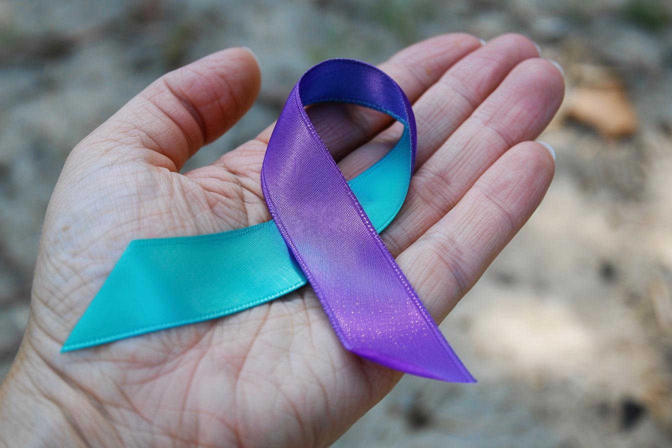 A hand holds a two-toned awareness ribbon with one half teal and the other half purple. The ribbon lies flat against the palm, with the background appearing blurred and out of focus.