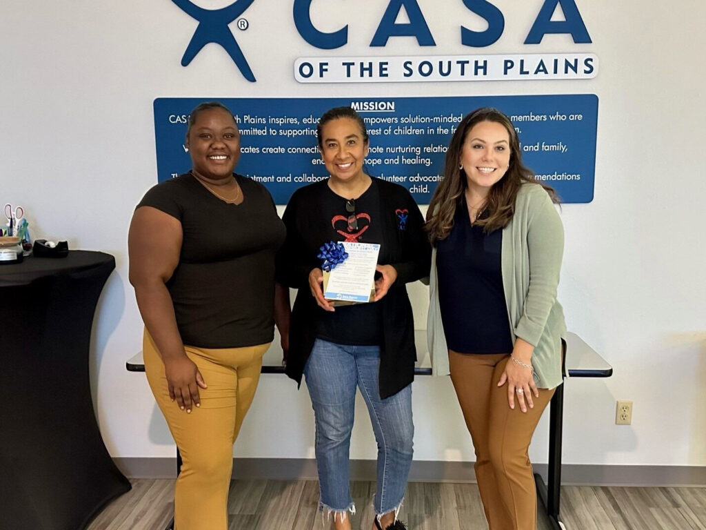 Three women are standing together in front of a wall with the CASA of the South Plains logo and mission statement. One of the women in the center is holding a small gift with a blue ribbon. They are smiling and appear to be at an informal event.