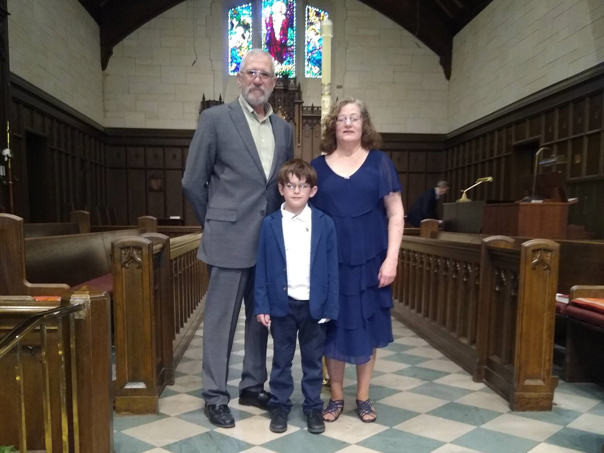 A man, woman, and young boy stand together inside a church. The man is wearing a gray suit, the woman a navy blue dress, and the boy a white shirt with a navy blazer and pants. Stained glass windows and wooden pews are visible in the background.