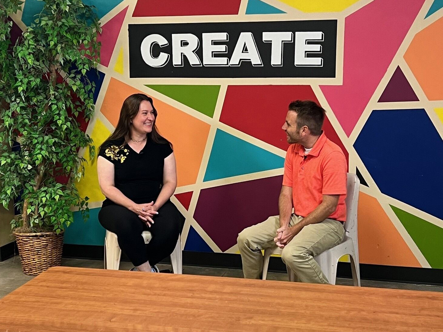 Two people sit on plastic chairs in front of a colorful wall with geometric patterns and the word "CREATE" displayed prominently. One person wears a black shirt, and the other wears an orange polo shirt. There is a potted plant to the left and a wooden table in the foreground.