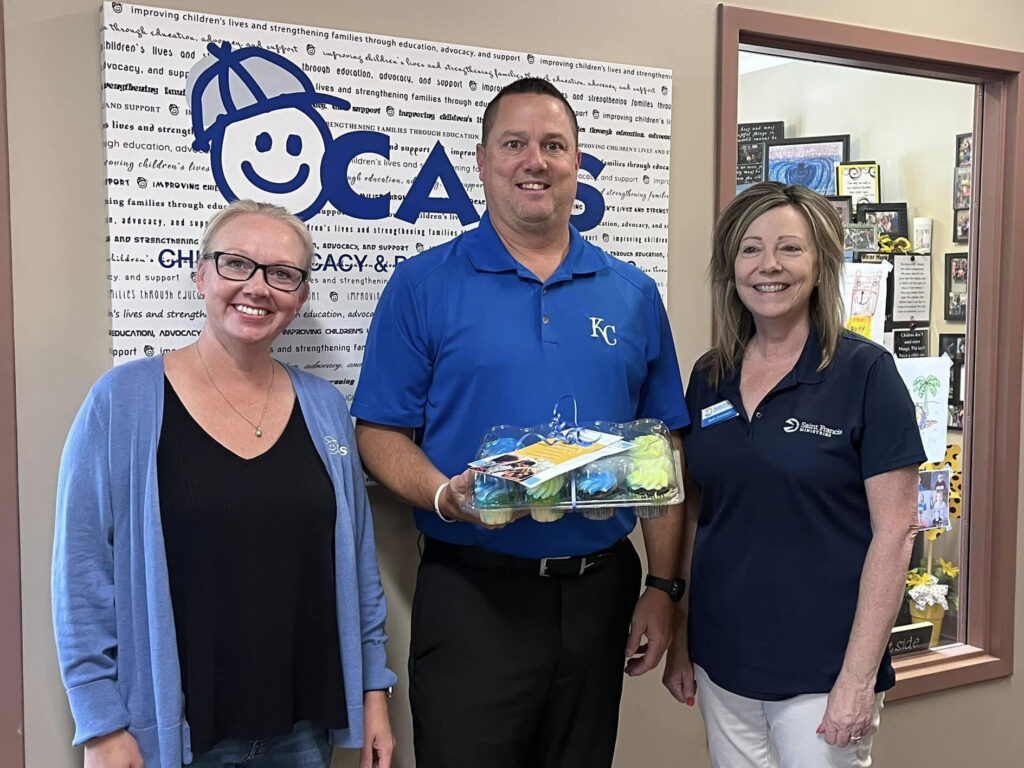 Three people are standing indoors, smiling in front of a sign with "CASA" written on it. The person in the center is holding a box of colorful cupcakes. They are all dressed in casual attire, with the two individuals on the left and right wearing name tags.