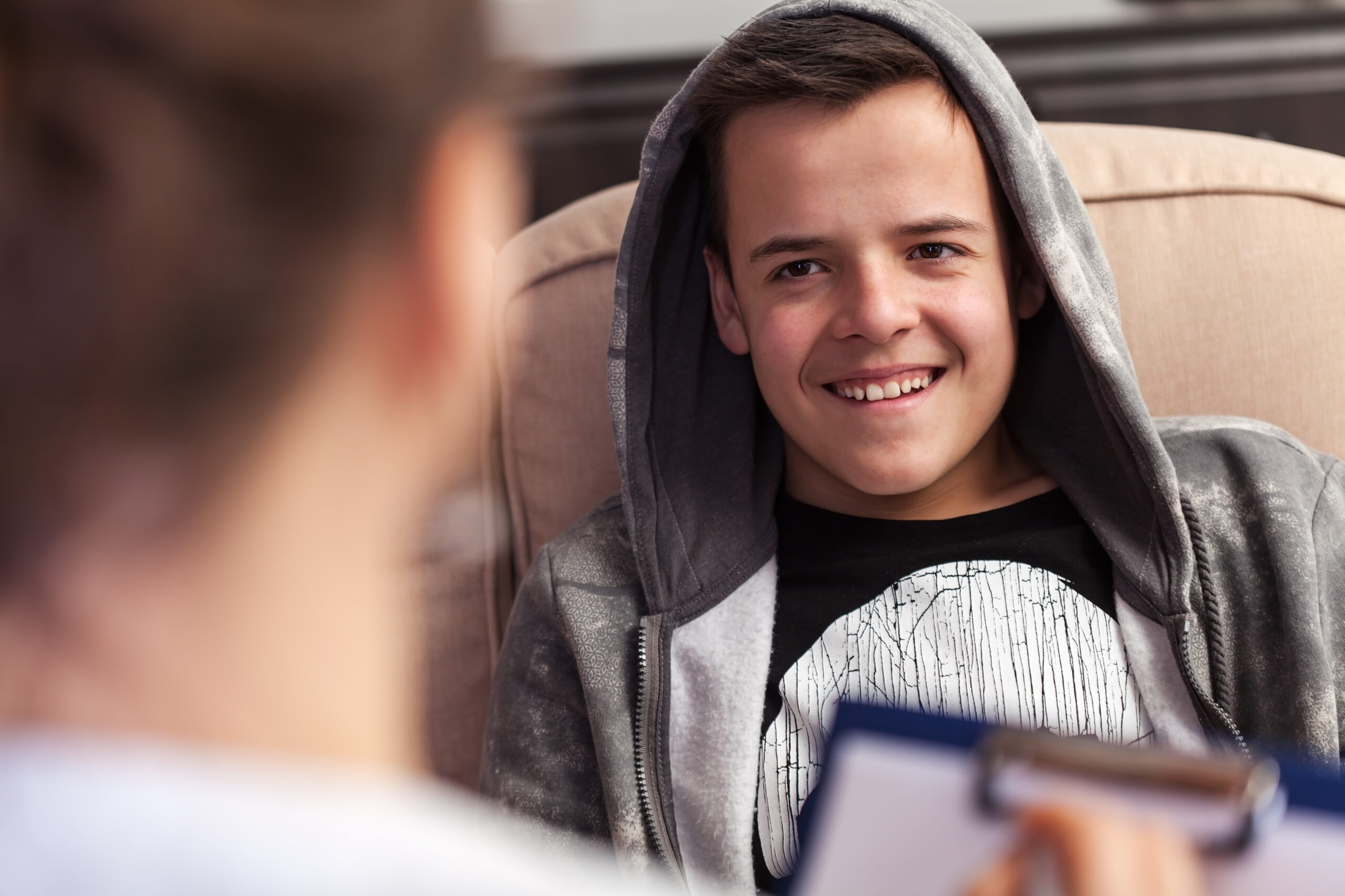 A teenage boy in a gray hoodie is sitting and smiling while looking at someone. He appears relaxed and engaged in conversation. The focus of the image is on him, with a blurred person holding a clipboard in the foreground.