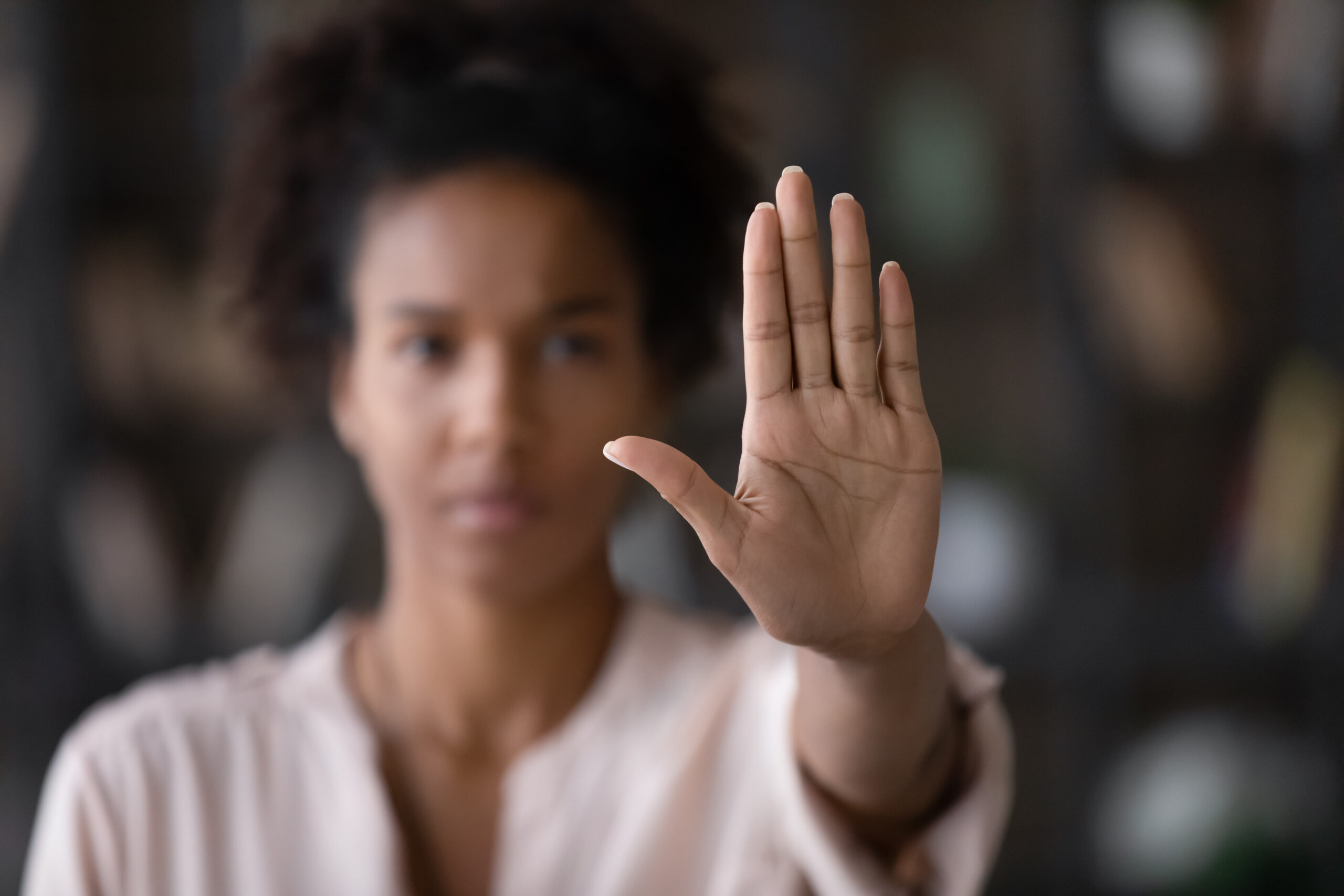 A person holding up their hand in front of them with fingers spread, as if gesturing to stop or block. The background is blurry, and the person is standing in a softly lit environment.