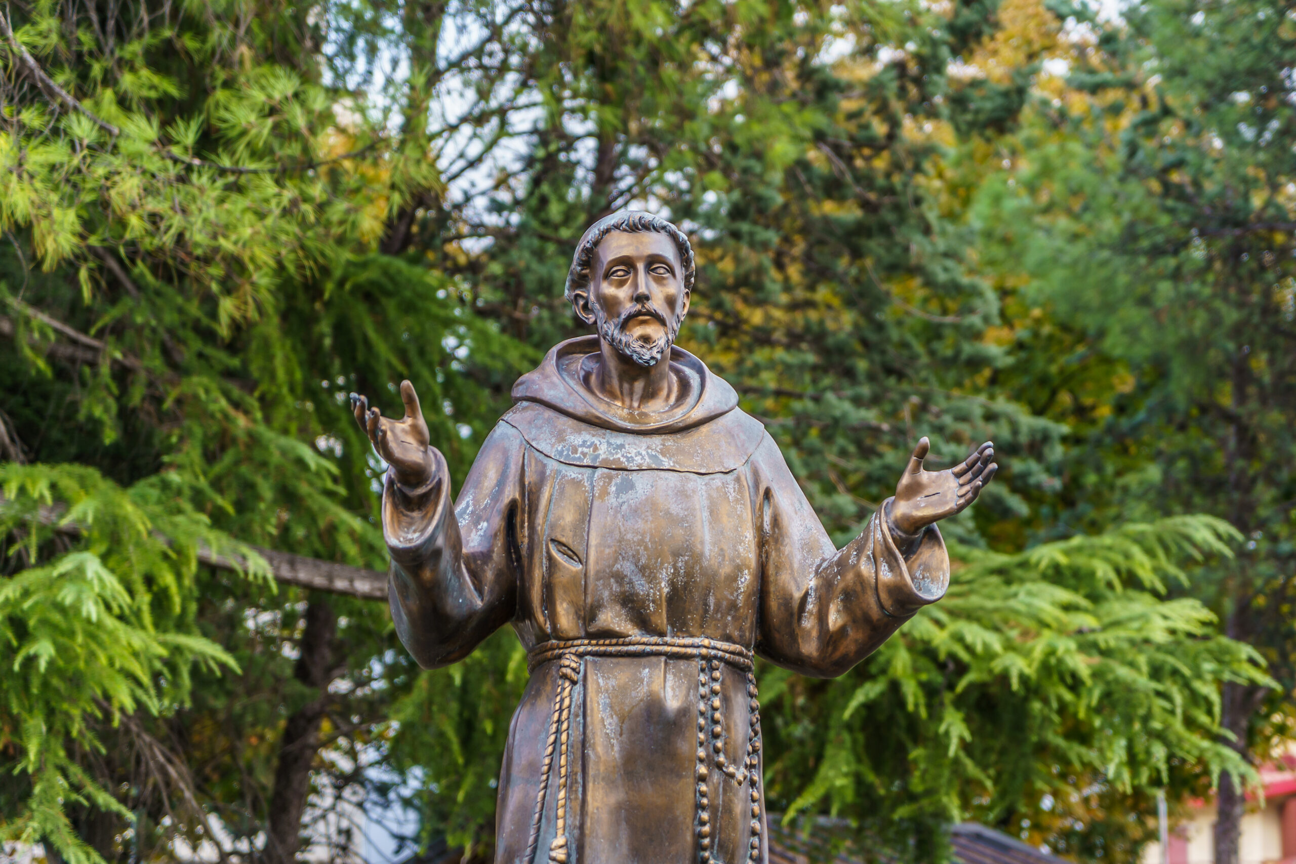 A bronze statue of a man wearing a robe with a rope belt, standing with both hands raised. The figure has a beard and a calm expression. The background features lush green trees.