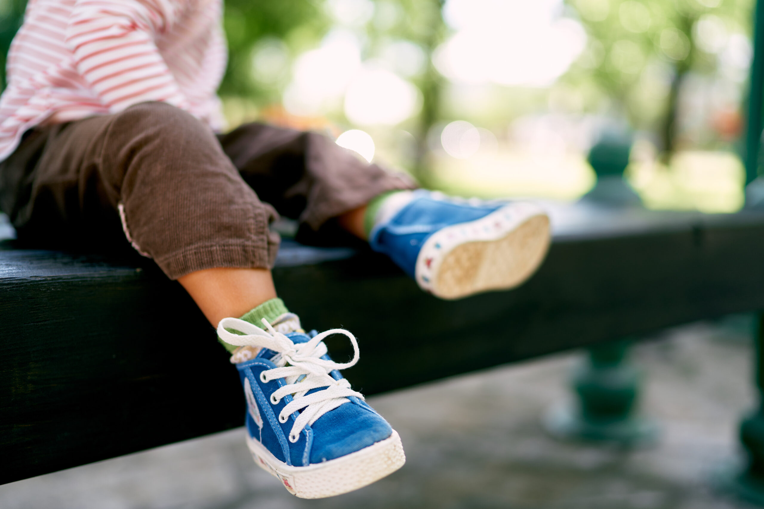 A child wearing blue sneakers and brown pants sits on a bench. The focus is on the shoes, which have white laces and rubber soles. The background is blurred with green trees and a bright, sunny atmosphere.