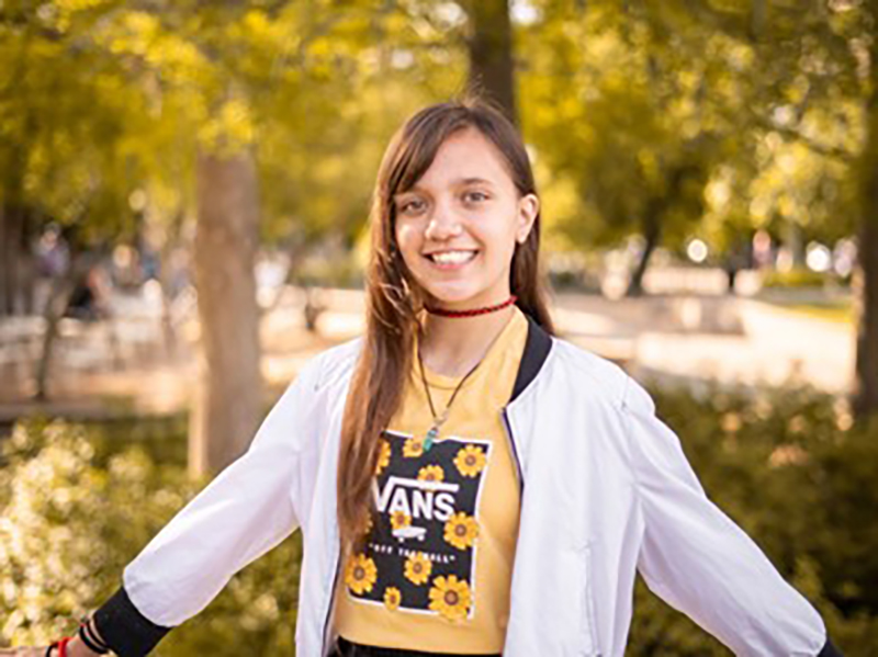 A young woman is standing outdoors with trees and greenery in the background. She is smiling, wearing a white jacket over a yellow crop top with a floral Vans logo, and has a red choker necklace. Her arms are slightly outstretched.