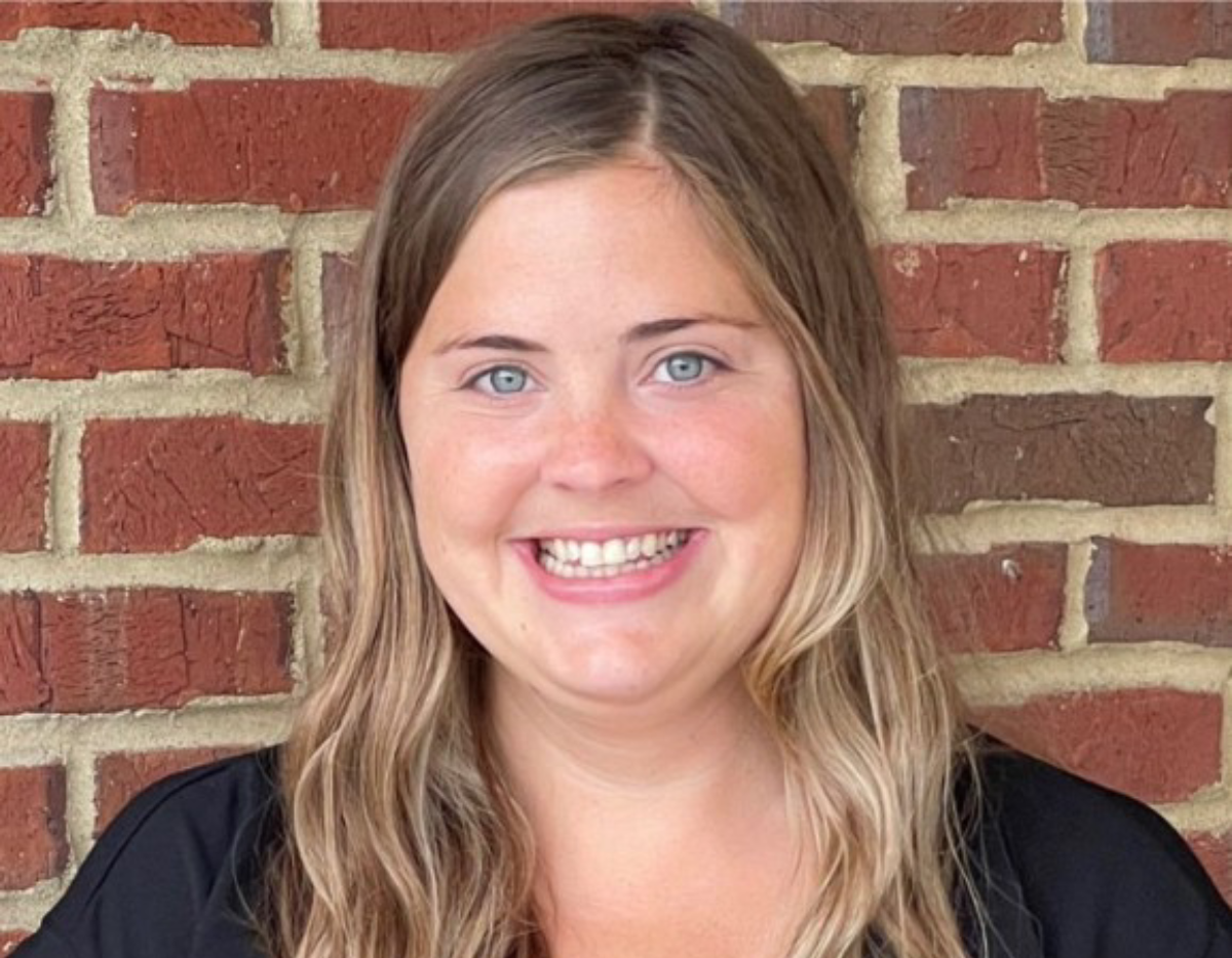 A person with long, light brown hair is smiling and facing the camera. They are standing in front of a red brick wall and wearing a black top.