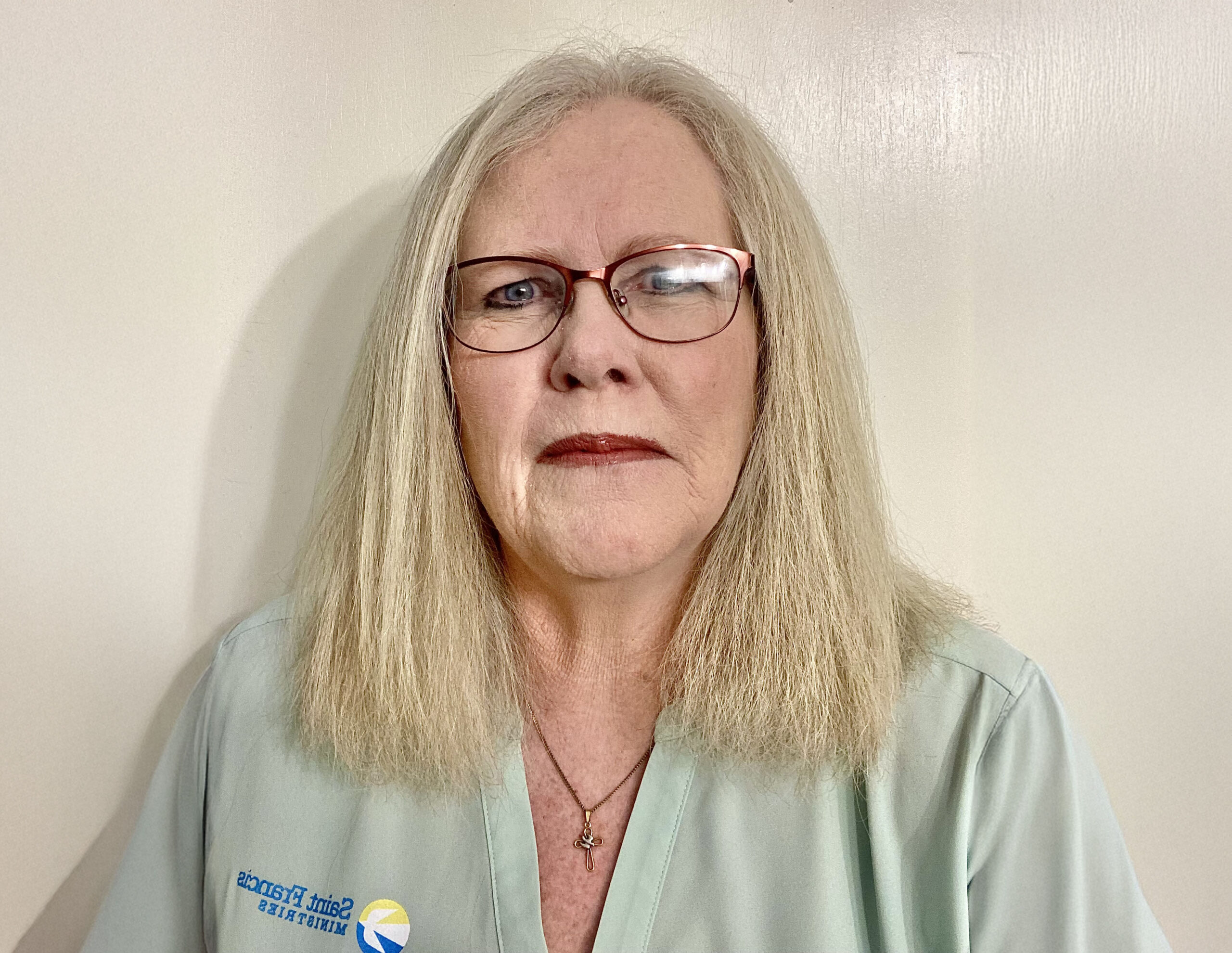 A person with long light hair and glasses is wearing a light blue-green shirt with a logo on it. They are standing against a plain light-colored background.