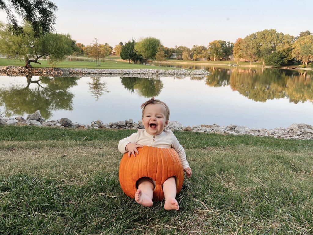 A baby sits inside a large pumpkin, laughing joyfully, with bare feet sticking out. The background features a calm lake, grassy park, and trees under a clear sky.