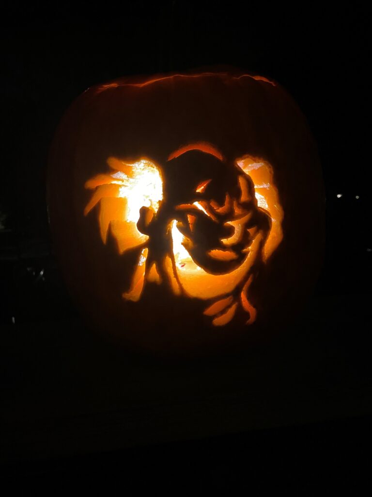 A carved pumpkin is lit from within, showcasing an intricate design of a stylized face with exaggerated features. The surrounding area is dark, highlighting the glowing effect of the carving.