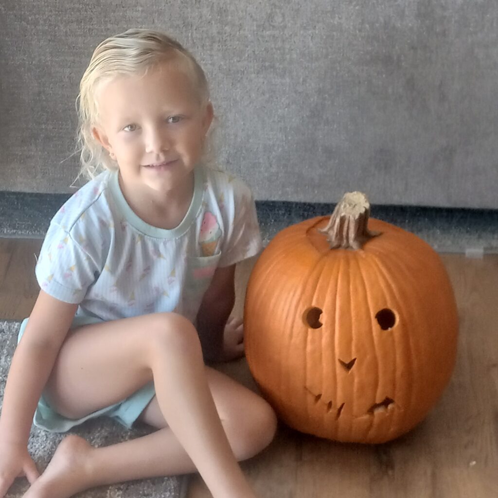 A child with short blond hair sits on a rug indoors next to a carved pumpkin. The pumpkin has a simple face design with triangular eyes and a jagged mouth. The child is wearing a light-colored shirt with a pattern.