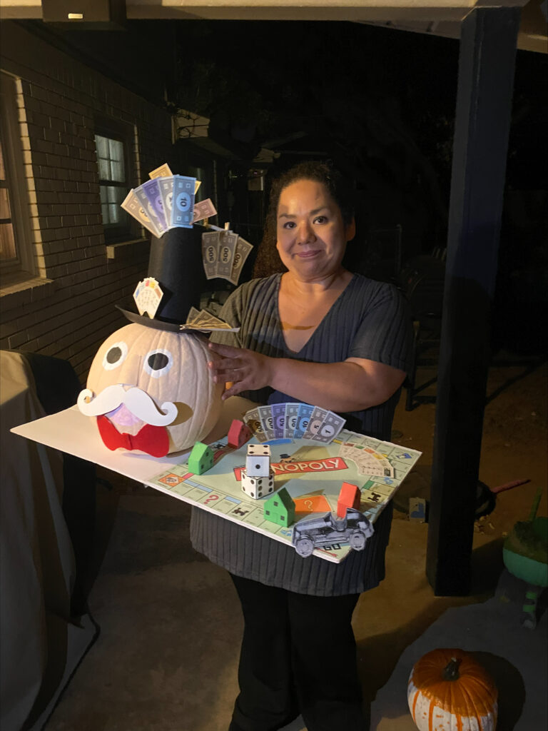 A person holding a decorated pumpkin resembling a Monopoly theme. The pumpkin features a top hat, mustache, and paper money. It rests on a board with Monopoly elements, including dice and hotels. A jack-o'-lantern is visible in the background.