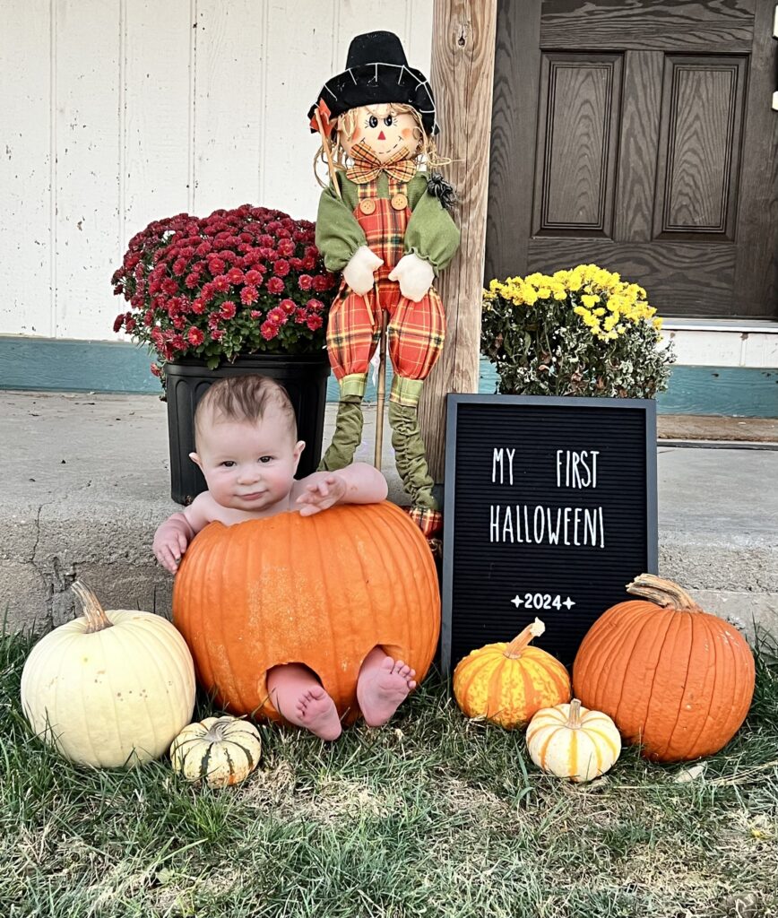 A baby sits inside a large orange pumpkin surrounded by smaller pumpkins and autumn flowers. A scarecrow decoration stands behind, and a letter board reads "My First Halloween 2024." The scene is set on a grass lawn in front of a porch.