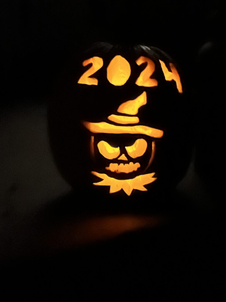 A carved pumpkin glows in the dark with the year "2024" at the top. Below, a design features a face with a hat and a sinister expression. Light emanates from the carved sections.