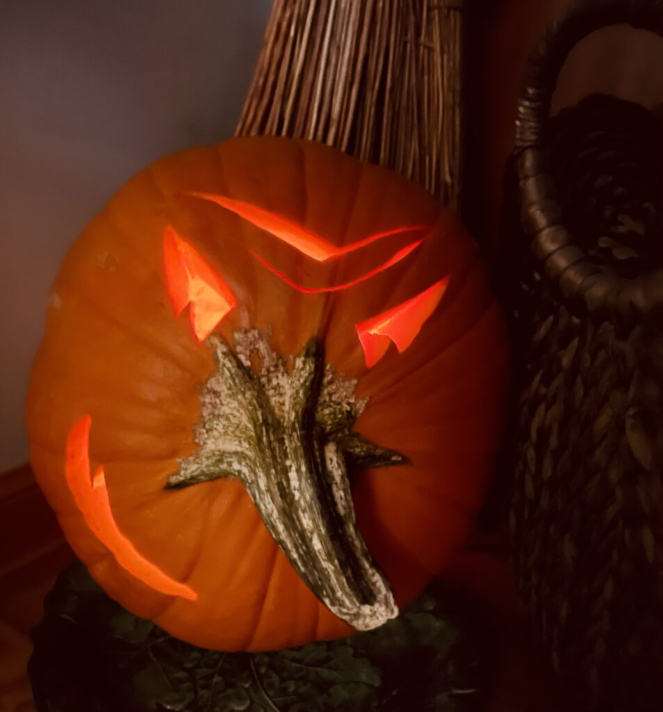 A carved pumpkin sits on a dark surface. It features an angry face design with glowing triangular eyes, eyebrows, and a jagged mouth illuminated from within. A white stem protrudes from the center. A large basket and broom are in the background.