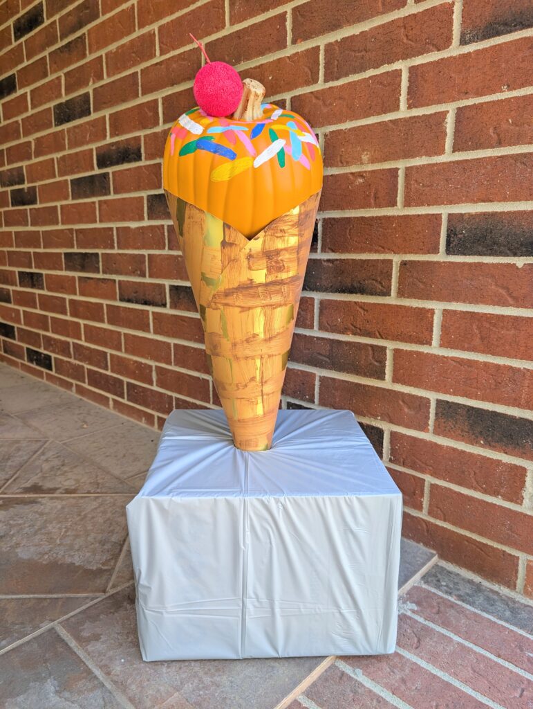 A sculpture of an ice cream cone sits on a wrapped pedestal. The cone is painted with a pattern resembling waffle texture, topped with bright colors and a red sphere representing a cherry. It is positioned in front of a brick wall.