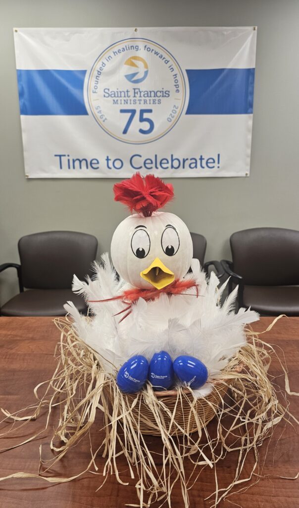 A craft resembling a white bird with a red bow and yellow beak sits on a nest made of straw and white feathers. Blue plastic eggs are in the nest. A banner behind reads "Saint Francis Ministries 75" and "Time to Celebrate!.