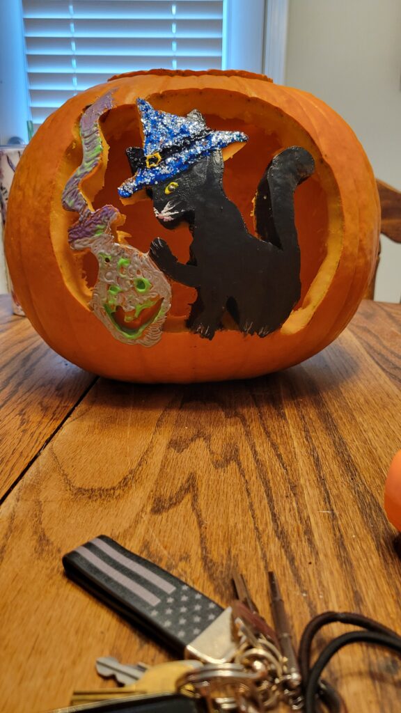 A carved pumpkin features a design of a black cat wearing a blue witch hat, with a green, spooky face next to it. The pumpkin is placed on a wooden table, and part of a keychain is visible in the foreground.