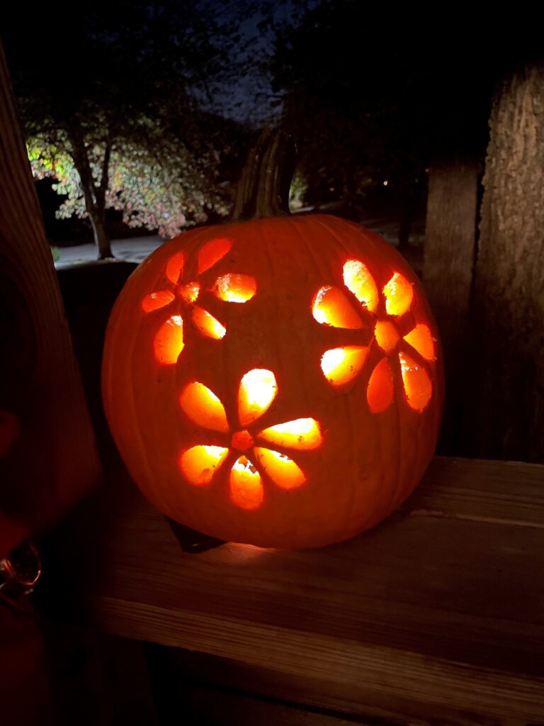 A carved pumpkin with illuminated flower patterns sits on a wooden surface. The background is dimly lit, with trees visible in the distance.