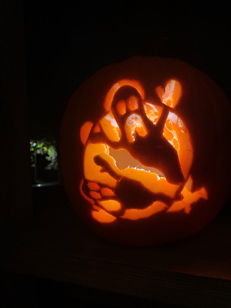 A lit jack-o'-lantern with a carved design of a ghost waving its hand. The pumpkin glows warmly in the dark, with intricate details visible in the carving.