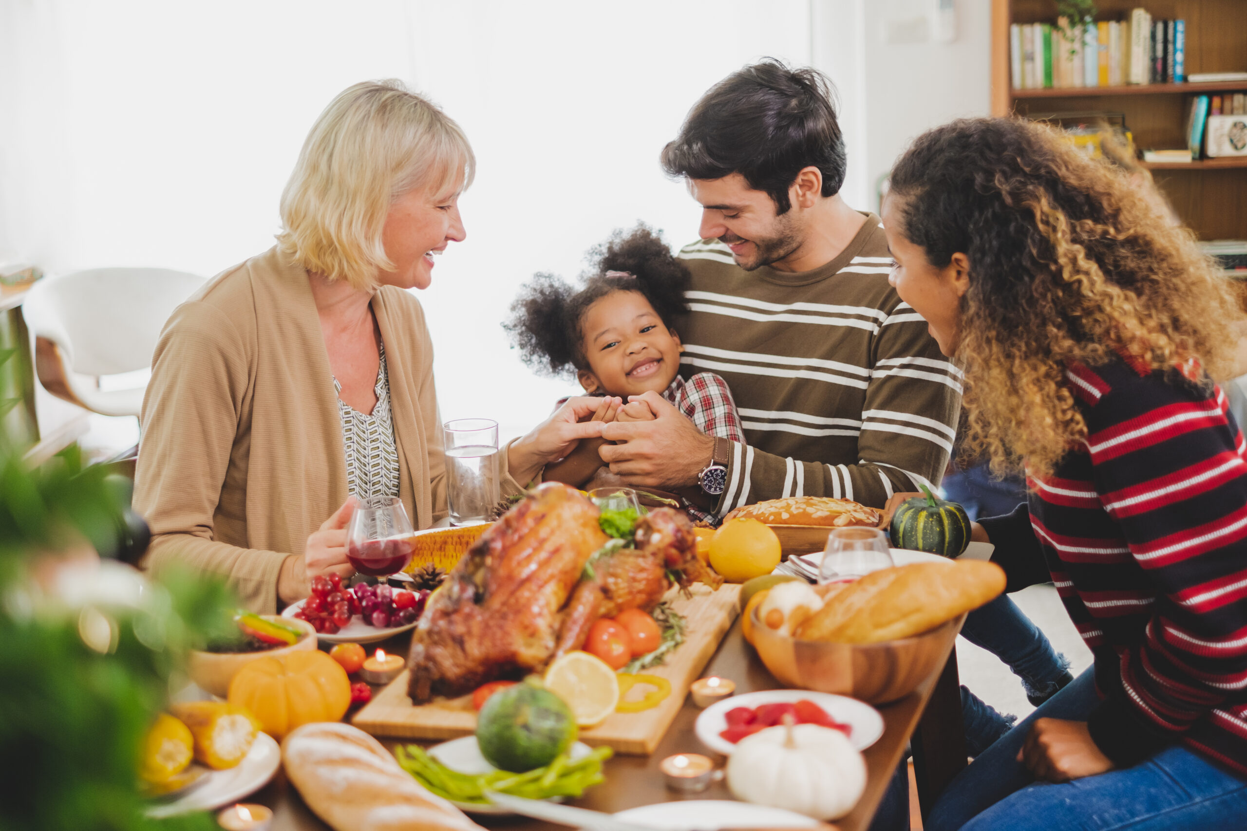 A family of four sits around a table, smiling and holding hands. The table is set with a roast turkey, candles, fruits, and bread. The setting appears warm and festive, with natural light coming through a window in the background.