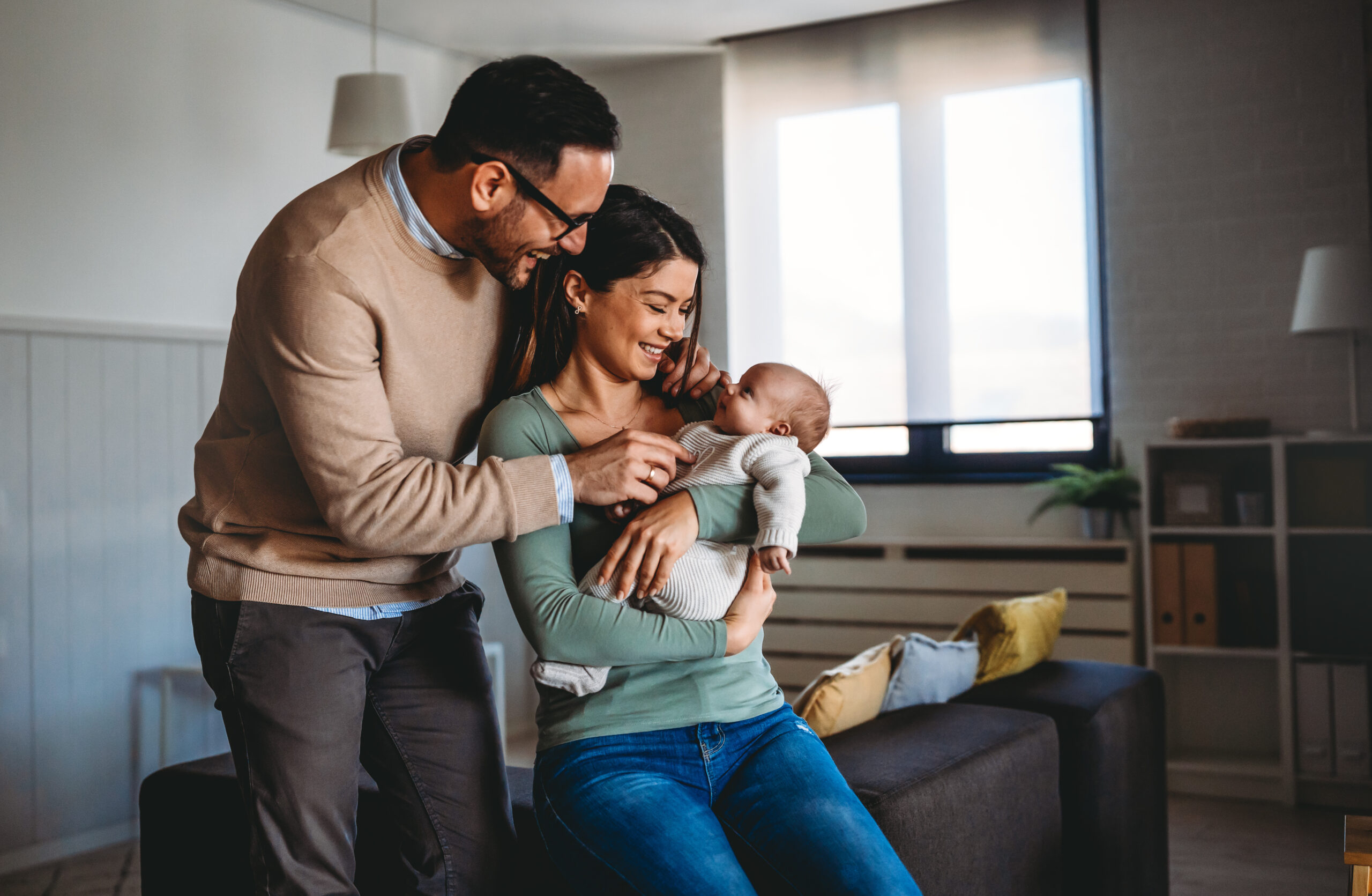 A man and a woman smile affectionately at a baby they are holding in a cozy living room. The woman is seated on a sofa, while the man leans over her. Soft lighting and simple decor create a warm atmosphere.