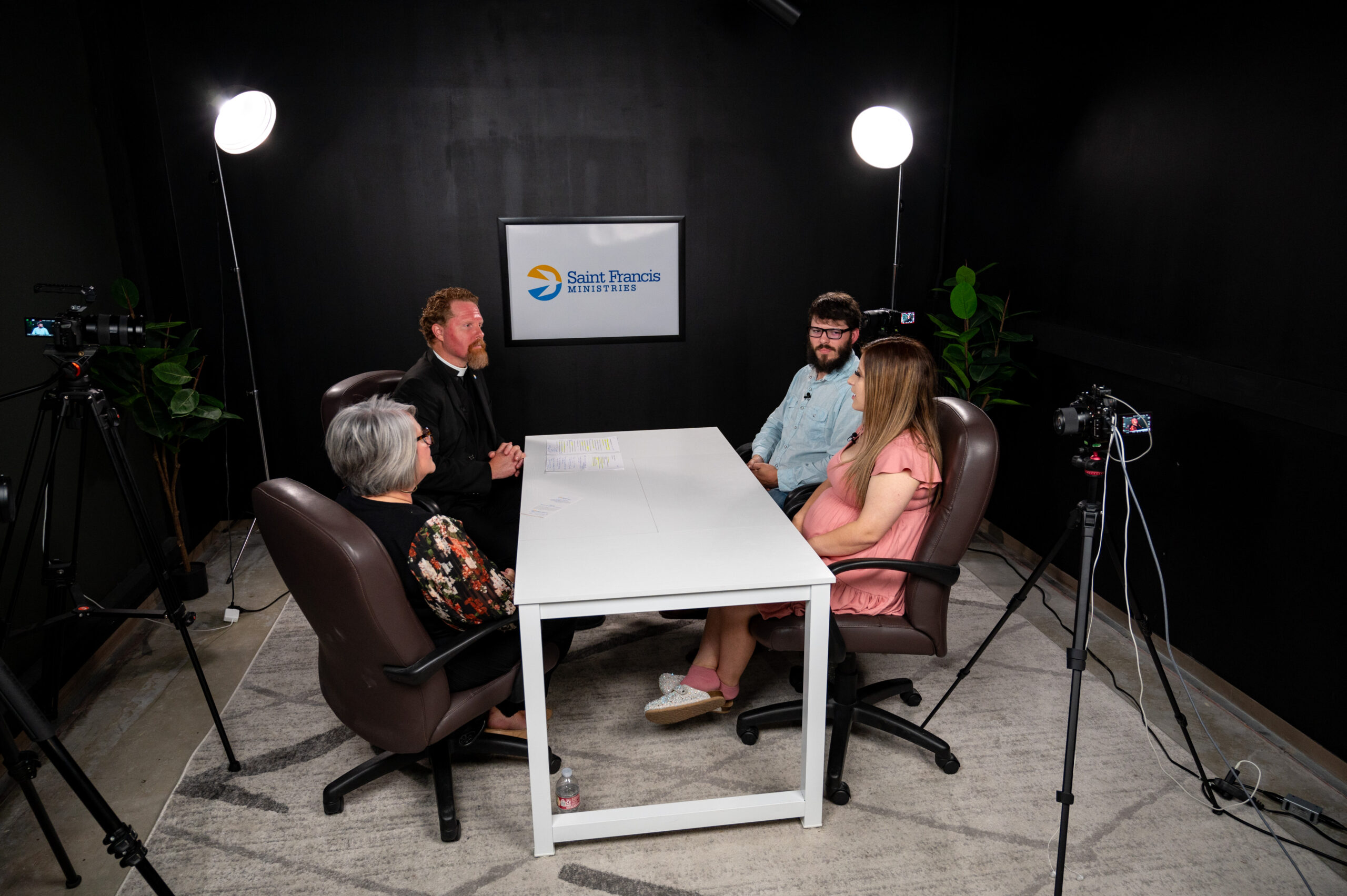 Four people sit around a white table in a dimly lit room with black walls and plants. Two men and two women face each other, and the table has recording equipment around it. A sign on the wall reads "Saint Francis Ministries.