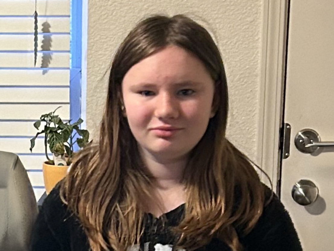 A young girl with long brown hair sits indoors near a window with closed blinds. A small potted plant is visible in the background alongside a door with a silver handle. She is wearing a black top.