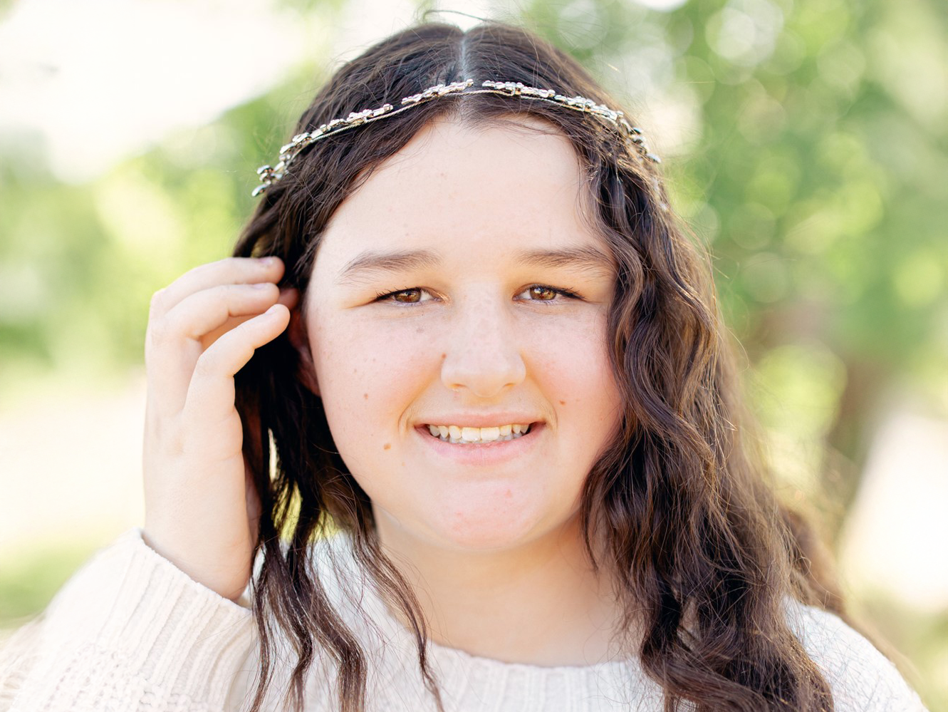 A person with long wavy hair, wearing a light sweater and a decorative headband, smiles at the camera. The background is blurred with green foliage.