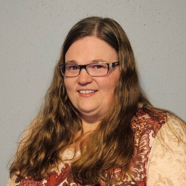 A woman with long brown hair and glasses is smiling. She is wearing a patterned blouse with shades of red and beige, standing against a plain light blue wall.