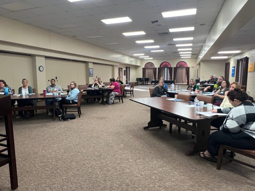 A group of people sitting at tables in a large, well-lit room, engaged in a meeting or presentation. Papers, water bottles, and other materials are on the tables. The room has a carpeted floor and neutral-colored walls.