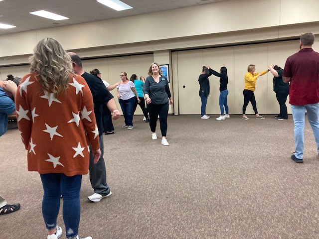 A group of people is participating in a dance class in a spacious room with beige walls and carpeted floors. Individuals are paired up, practicing dance moves. A woman in a green shirt walks towards the camera, smiling.