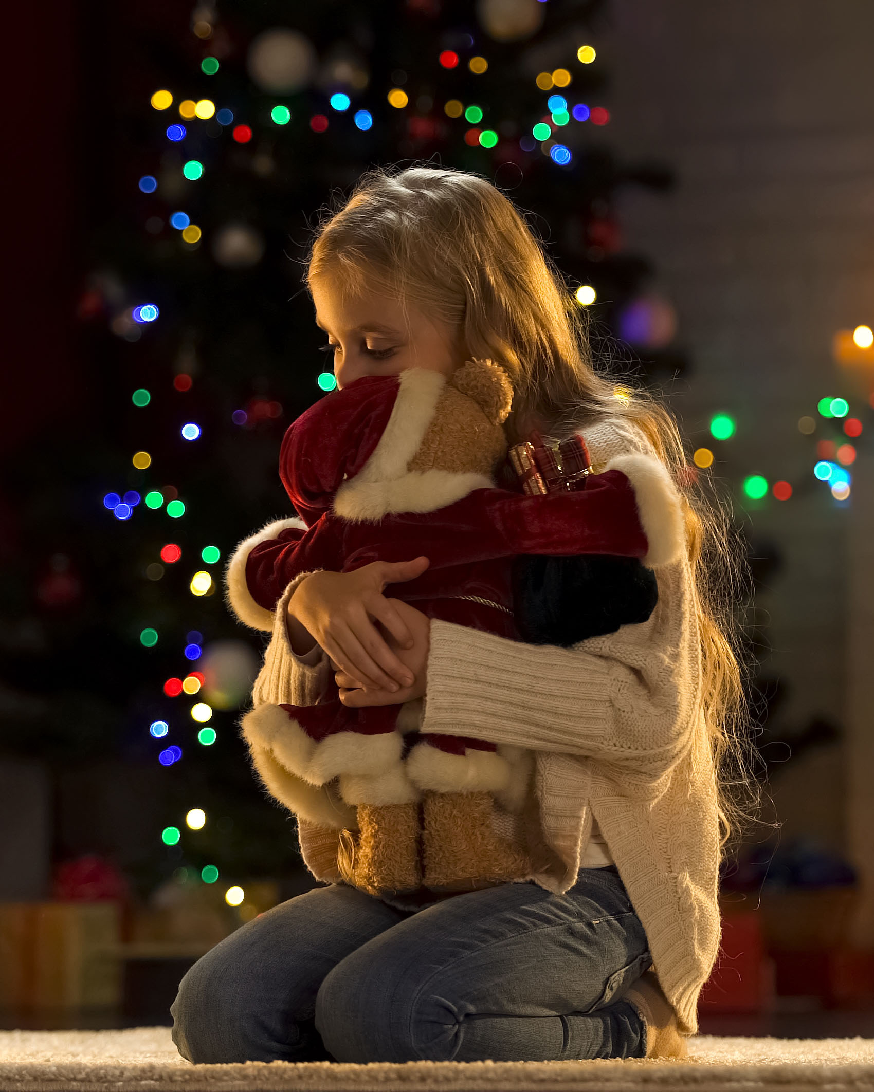 A child with long hair, wearing a sweater and jeans, is kneeling on the floor, hugging a teddy bear dressed in a Santa outfit. In the background, a Christmas tree is decorated with colorful lights.