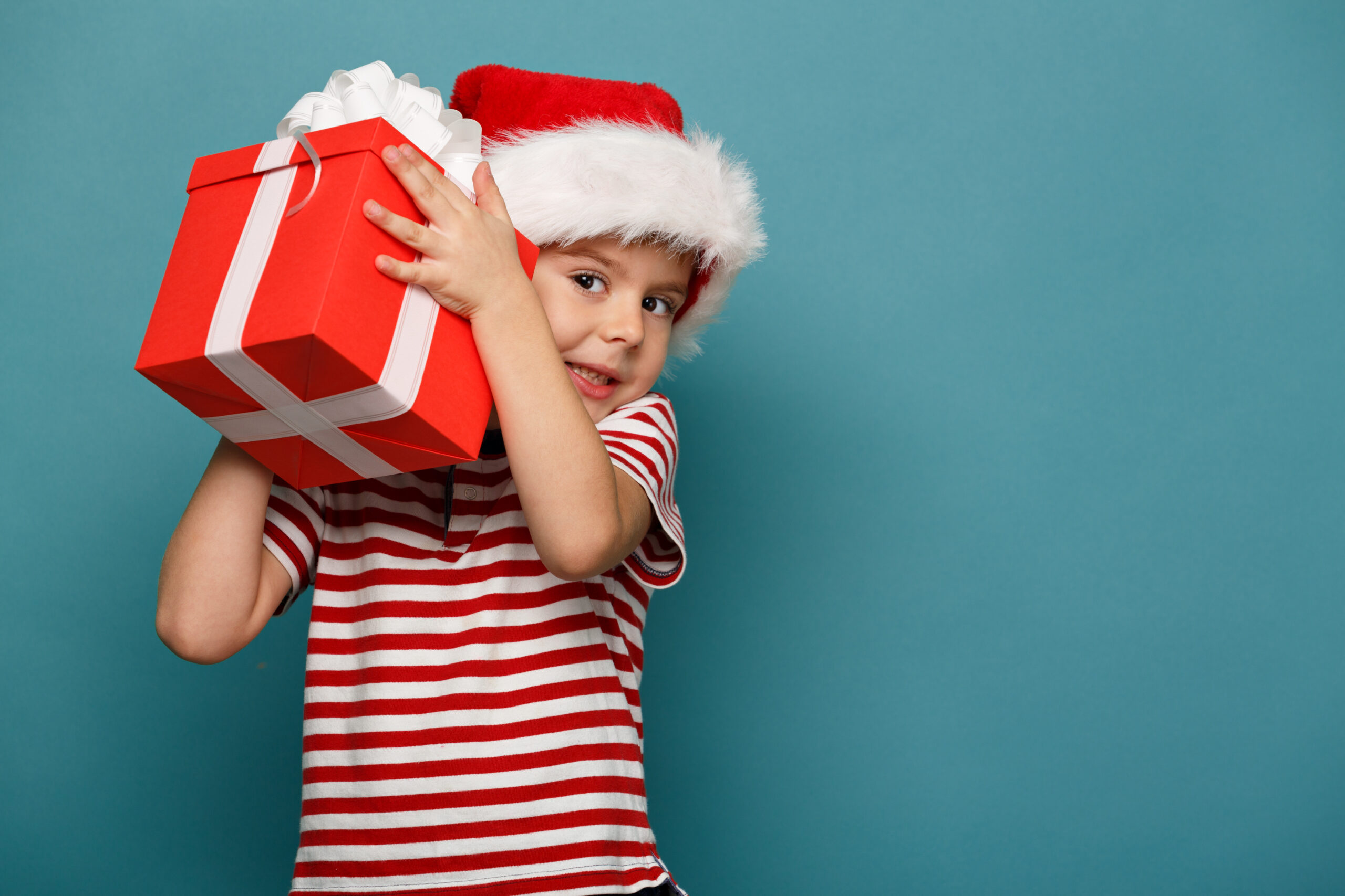 A child wearing a Santa hat and a red and white striped shirt holds a red gift box with a white ribbon against a teal background.