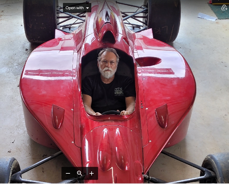 A man with a beard is seated in a shiny red racing car in a garage setting, looking directly at the camera. The car has a sleek design and the overhead view shows its aerodynamic shape.