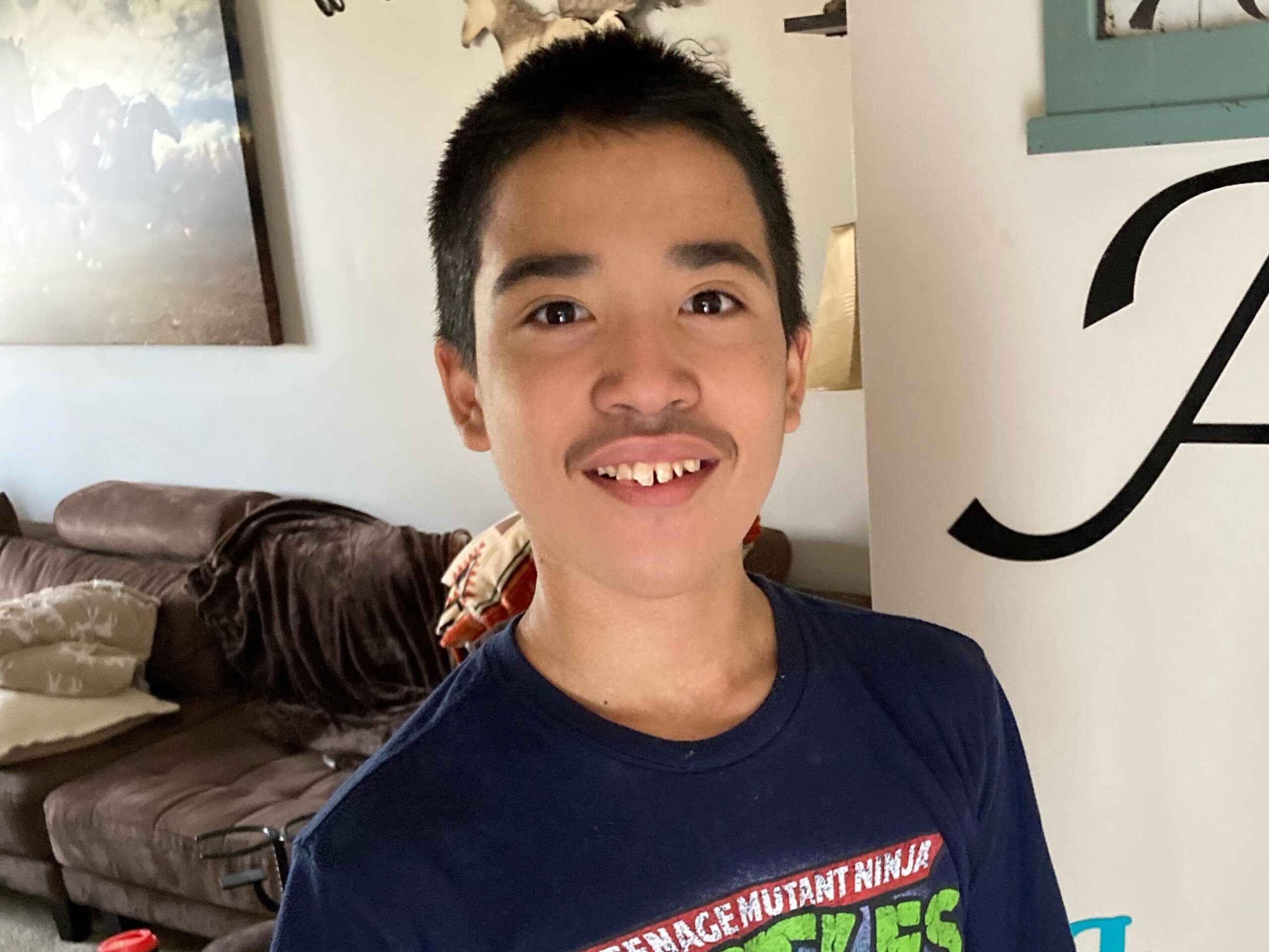 A young person with short dark hair and a mustache smiles at the camera. They are wearing a Teenage Mutant Ninja Turtles shirt. Behind them is a brown couch with pillows and a wall with framed art.