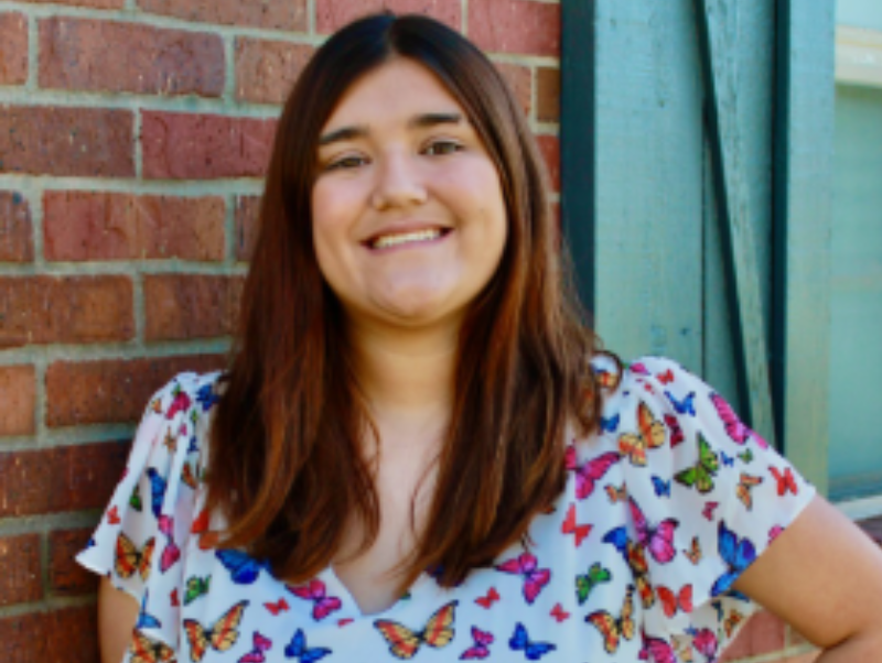 A person with long brown hair is smiling, standing against a brick wall. They are wearing a white shirt with a colorful butterfly pattern. The background includes part of a wooden window frame.