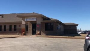 A single-story brick and beige building with a sign reading "Starr Home Health" above the entrance. The building is surrounded by a concrete driveway and a clear blue sky overhead. Cars are partially visible on the right side.