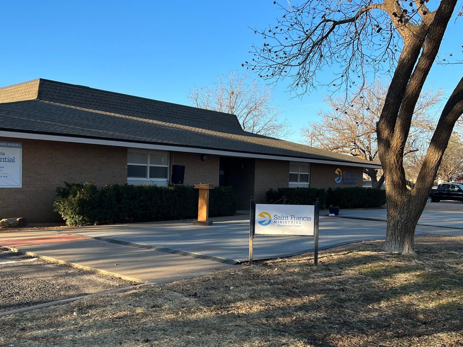 Single-story brick building with a sign reading "Saint Francis Ministries" in front. A bare tree and shrubbery are present. The sky is clear, and the ground is covered with dry grass and concrete paths.