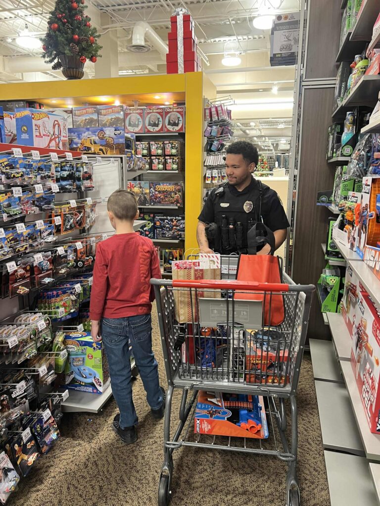 A child in a red shirt stands in the toy aisle of a store, looking at items on the shelf. A uniformed police officer with a shopping cart filled with toys stands next to him. The aisle is filled with various toys and games.